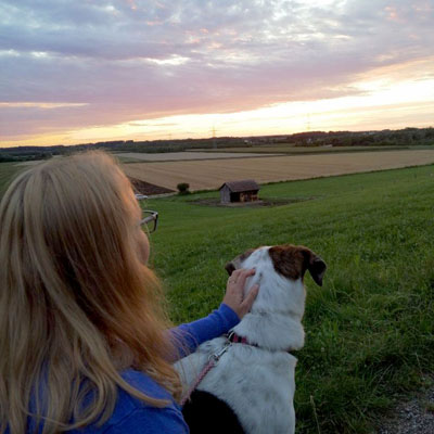 Links unten im Bild ist Alwina Simon von hinten zu sehen, wie sie mit der Hündin Lilly in Richtung Sonnenuntergang blickt. Dabei hat sie ihre linke Hand auf dem Kopf der Hündin abgelegt und streichelt sie. Der Blick geht hinunter ins Tal, über grüne Wiesen, okafarbene Felder und eine kleine Hütte. Am Horizont geht währenddessen die Sonne in gleißendem Licht unter und färbt die Wolkenformation darüber in einem milden Verlauf von Hellgelb über Zartrosa bis hin zu Hellblau. Bildtitel „Das bin ich mit Hündin Lilly“. / Das Bild stammt aus dem Jahr 2021 und ist Teil des privaten Bildarchivs von Alwina Simon.