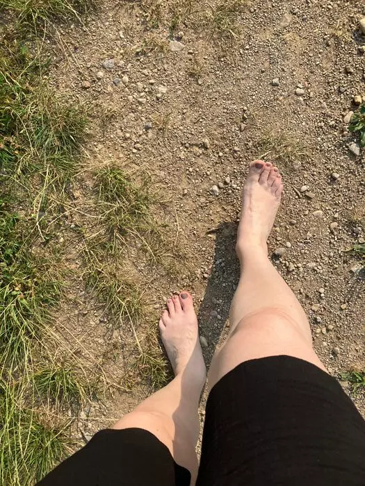 Blick von oben auf zwei nackte Füße, die barfuß über einen sonnigen Naturweg aus Erde und feinem Kies neben trockenem Gras gehen. Barfuß gehen auf einem Silent Walk.