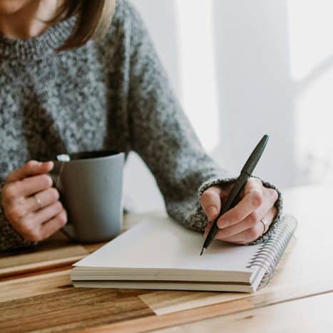 Frau sitzt an einem Holztisch, schreibt in ein Notizbuch und hält eine Tasse Kaffee in der Hand