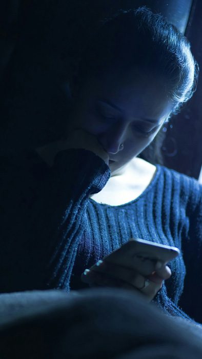 Frau sitzt abends im Dunkeln mit dem Handy, Sinnbild für Körperlosigkeit als Flucht aus dem Alltagsstress.