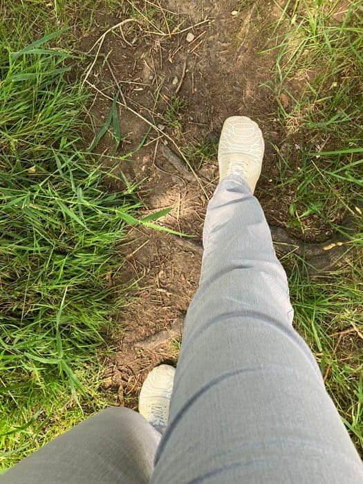 Silent Walk in der Natur, Blick von oben auf die eigenen Beine und hellen Barfußschuhe auf einem Erdweg zwischen grünem Gras