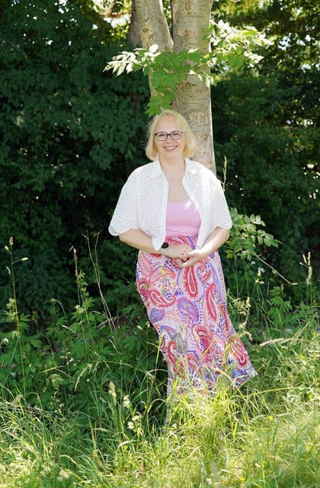 Alwina Simon steht lächelnd an einen Baum gelehnt in einer grünen, sommerlichen Wiese, in weißer Bluse über rosa Top und gemustertem Rock.