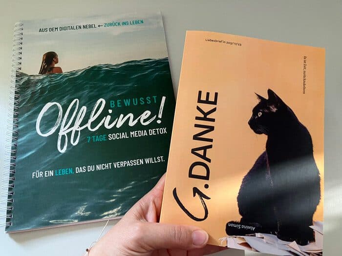 Das spiralgebundene Workbook „Bewusst Offline! 7 Tage Social Media Detox“ mit einer Surferin auf einer grünen Welle und das Buch „G.DANKE“ mit einer schwarzen Katze auf orangem Grund, beide von Alwina Simon, in einer Hand gehalten.