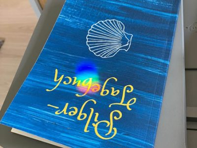 Beitragsbild der Podcastfolge_O-3 / Kreative Schöpfung, die sich in der realen Welt manifestiert _ Offen Gesprochen... über Gott und die Welt mit Martin Kolodziej und Alwina Simon - Ich & DU, Martin - Wow Sessions, ein Podcast über das Leben an sich // Zu sehen ist ein Schnappschuss eines auf dem Kopf liegenden blauen Pilgertagebuchs mit einer weißen Muschel als Umriss und dem Untertitel „Der Weg ist das Ziel“. Über dem „b“, das durch die 180-Grad-Drehung als „g“ zu lesen ist, liegt eine regenbogenfarbene Reflexion. // Credits: Privater Bildbestand von Alwina Simon – 2024-04-26