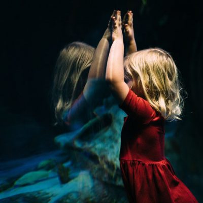 Ein kleines blondes Mädchen in einem roten Kleid steht an der Scheibe eines großen Aquariums. Ihre schemenhafte Reflexion ist in dem Glas erkennbar. Sie hat die Arme nach oben auf das Glas gelegt. Es sieht so aus, als berühre sie einen geisterhaften Zwilling, der sich hinter der Scheibe befindet. / Credits: Bekah Russom via unsplash.com