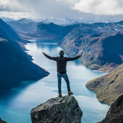 Auf einem Felsen stehend ist ein Mann von hinten in Ganzkörperansicht zu sehen, der die Arme ausbreitet. Vor ihm erstreckt sich der Blick auf einen von hohen Bergen umschlossenen Bergsee. Am Himmel sind weiße Wolken zu sehen. / Credits: Julian Hanslmaier via unsplash.com