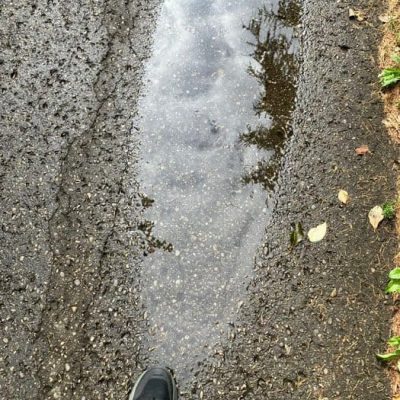 Pfütze auf einem Weg spiegelt den bewölkten Himmel und einen Baum, davor die Füße der Spaziergängerin Alwina Simon, Sinnbild für achtsam spazieren und sehen, was IST.