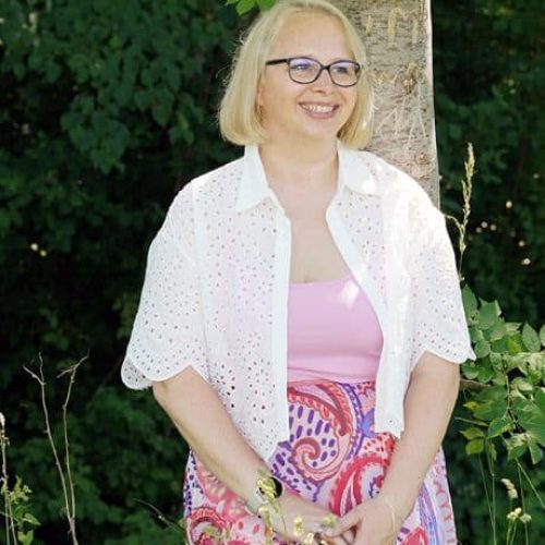 Alwina Simon steht lächelnd an einen Baum gelehnt in einer grünen, sommerlichen Wiese, in weißer Bluse über rosa Top und gemustertem Rock, Portrait.