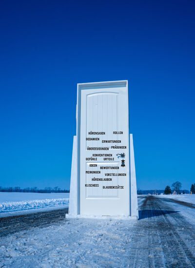 Im Freien, in einer Winterlandschaft, steht eine helle, bläulich schimmernde Holztür. Sie ist von einem Rahmen gehalten und befindet sich mitten auf einer Straße mit Fahrspuren. Im Hintergrund strahlt ein wolkenfreier, leuchtend blauer Himmel. Auf der Tür befinden sich Begriffe (Hörensagen, Rollen, Gedanken, Erwartungen, Prägungen, Überzeugungen, Konventionen, Gefühle, Urteile, Ideen, Bewertungen, Meinungen, Vorstellungen, Hörenglauben, Klischees, Glaubenssätze). / Credits: Topsphere Media via unsplash.com