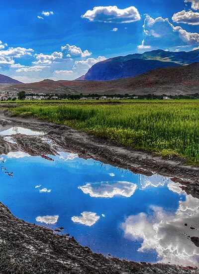 Auf schlammigem Boden steht eine große Pfütze, in der sich die Wolken des Himmels spiegeln. Das Bild ist sehr kontrastreich und zeigt intensive Blau- und Grüntöne. / Credits: Towhid Shamsi via unsplash.com